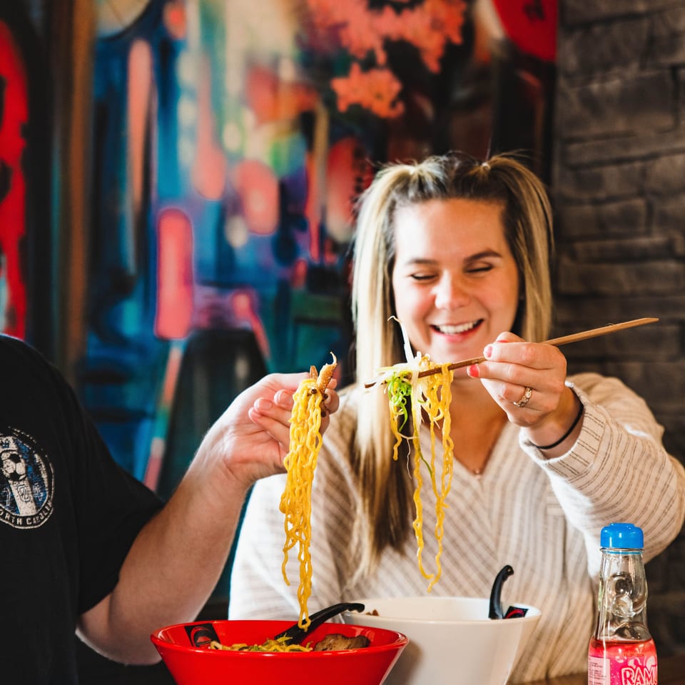 Smiling diners lift noodles with chopsticks over red and white bowls in a colorful restaurant.