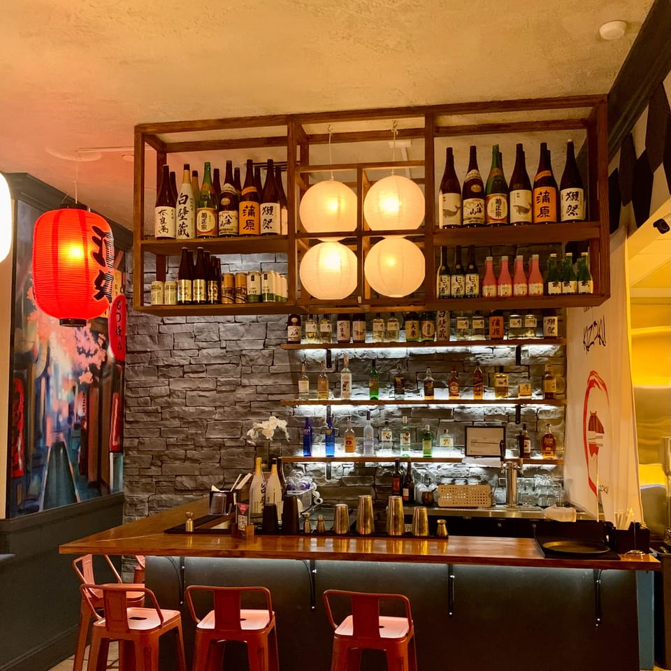Warmly lit Japanese restaurant bar with shelves of sake and spirits, paper lanterns, and red stools.