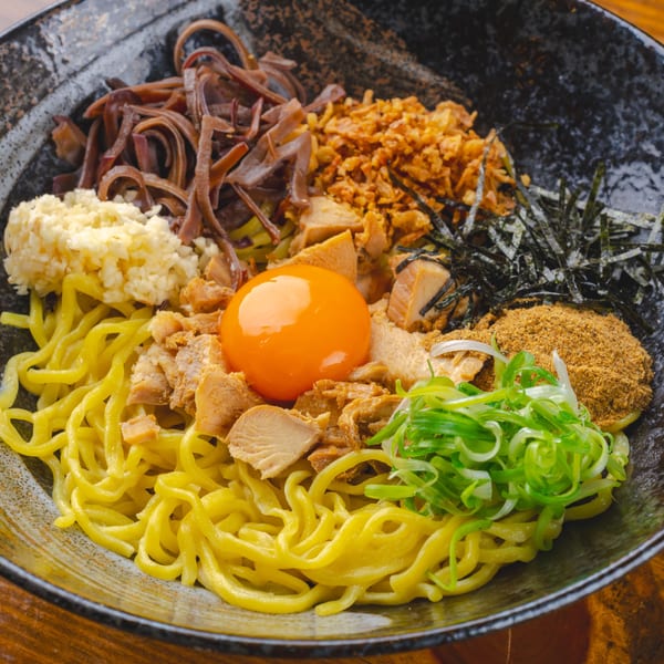 A bowl of mazesoba with noodles, pork, egg yolk, green onions, and other toppings.