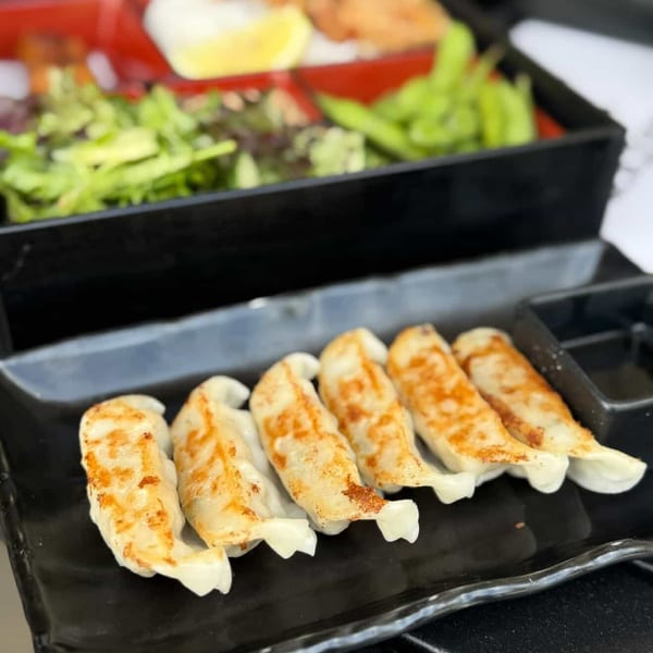 Pan-fried gyoza with a bento box of chicken, rice, salad, and edamame in the background.