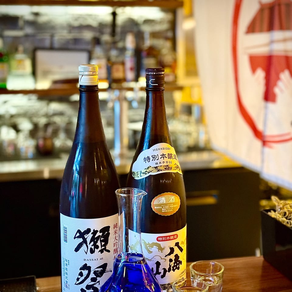 Japanese sake bottles, blue serving carafe, and glasses arranged on a wooden restaurant table.