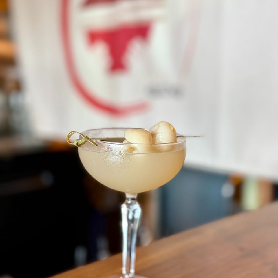 Cream-colored cocktail in a stemmed glass, garnished with fruit and a skewer, sits on a wooden bar.