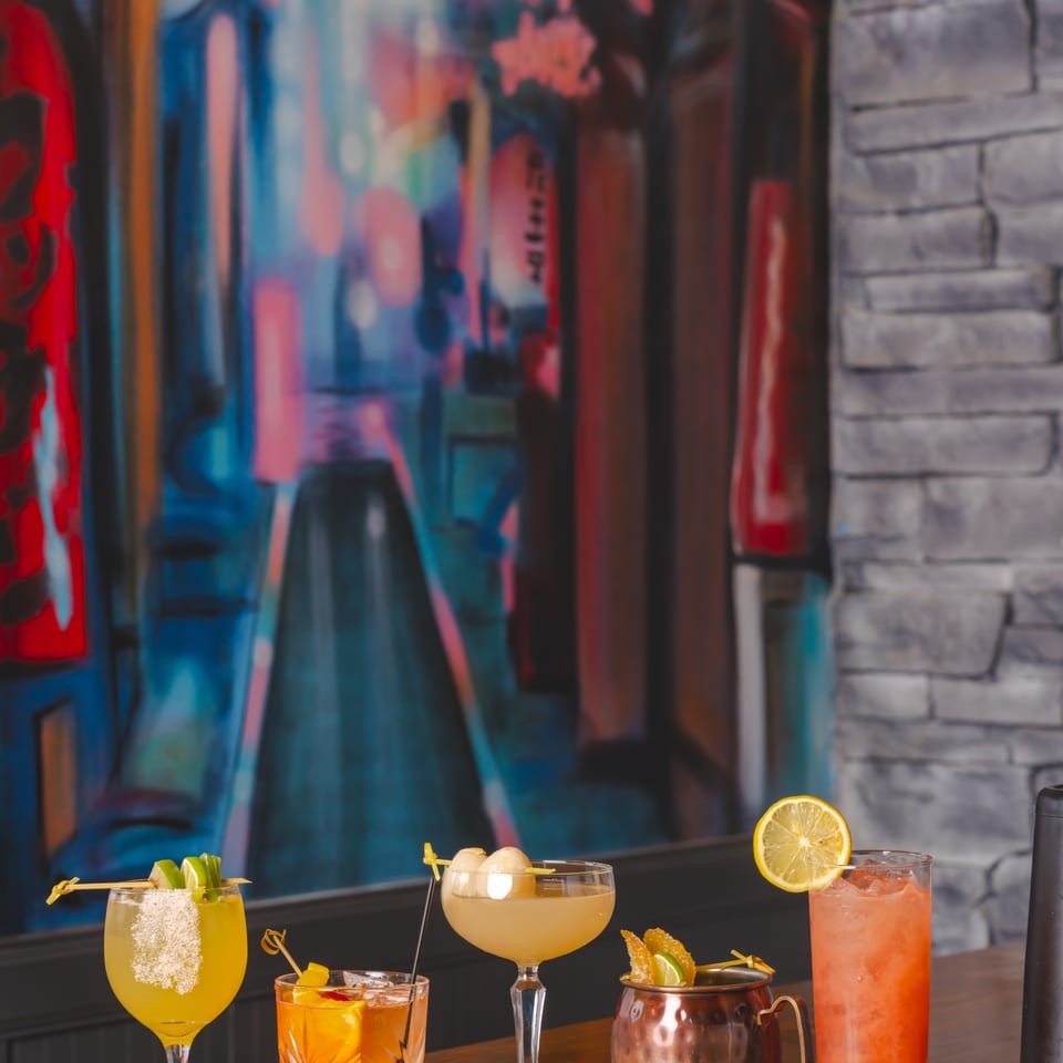 Colorful cocktails garnished with citrus and fruit sit on a polished wooden bar against a vibrant mural backdrop.
