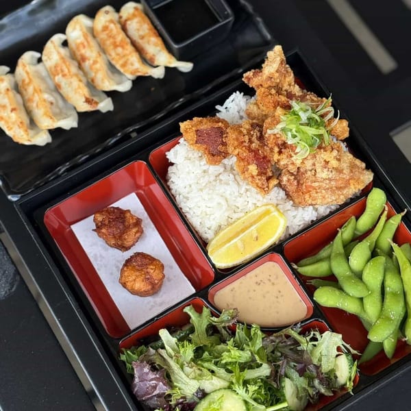 A bento box with fried chicken, rice, lemon, edamame, salad, dipping sauce, and gyoza.