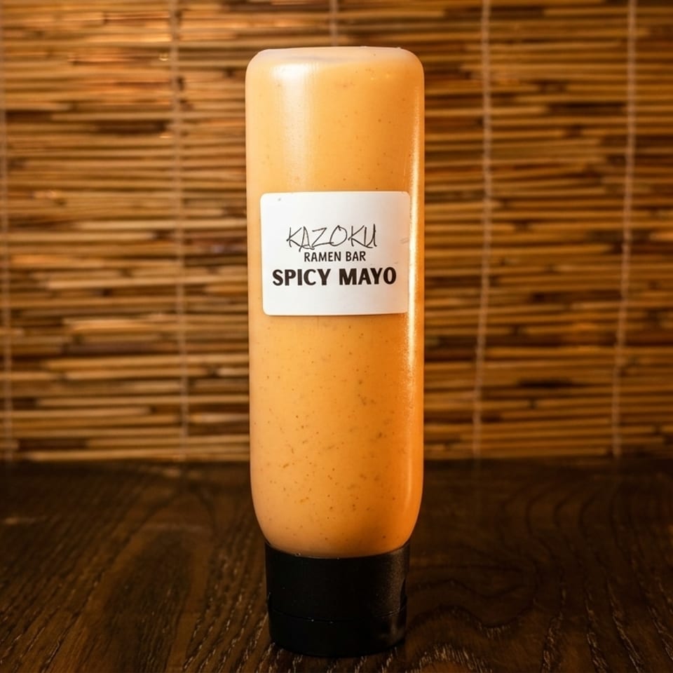 A bottle of Kazoku Ramen Bar Spicy Mayo stands on a dark wooden surface against a bamboo background.
