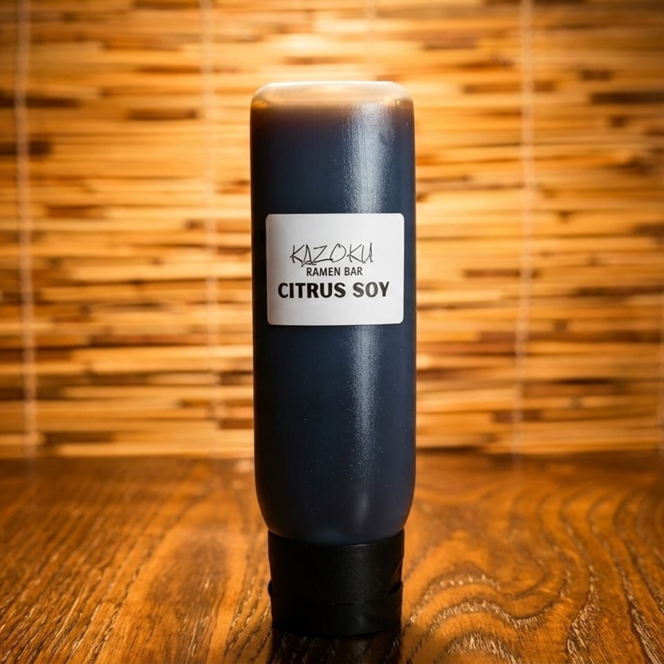 A bottle of Kazoku Ramen Bar Citrus Soy sauce on a wooden table with a bamboo mat background.