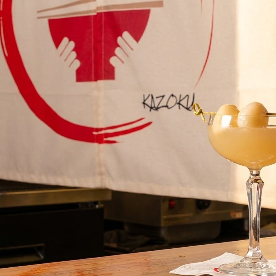 A light-colored cocktail with two lychee fruits on a pick, in front of a Kazoku banner.