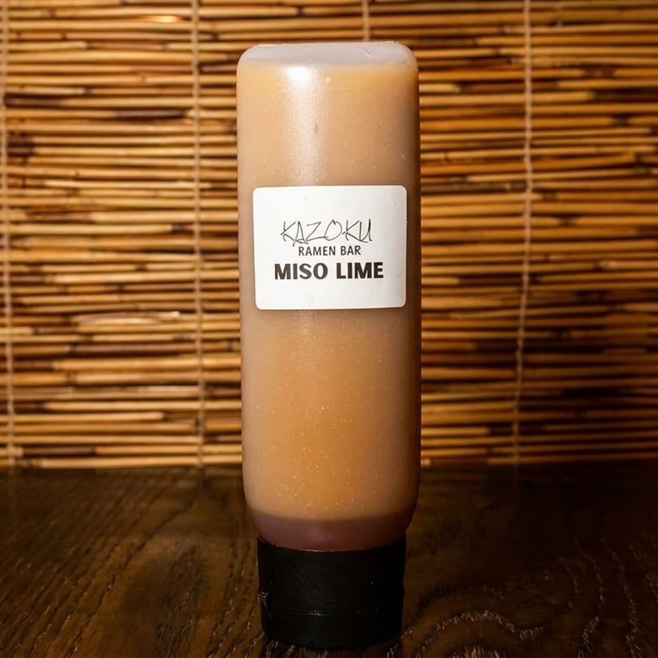 A squeeze bottle of Kazoku Ramen Bar Miso Lime sauce sits on a wooden table.